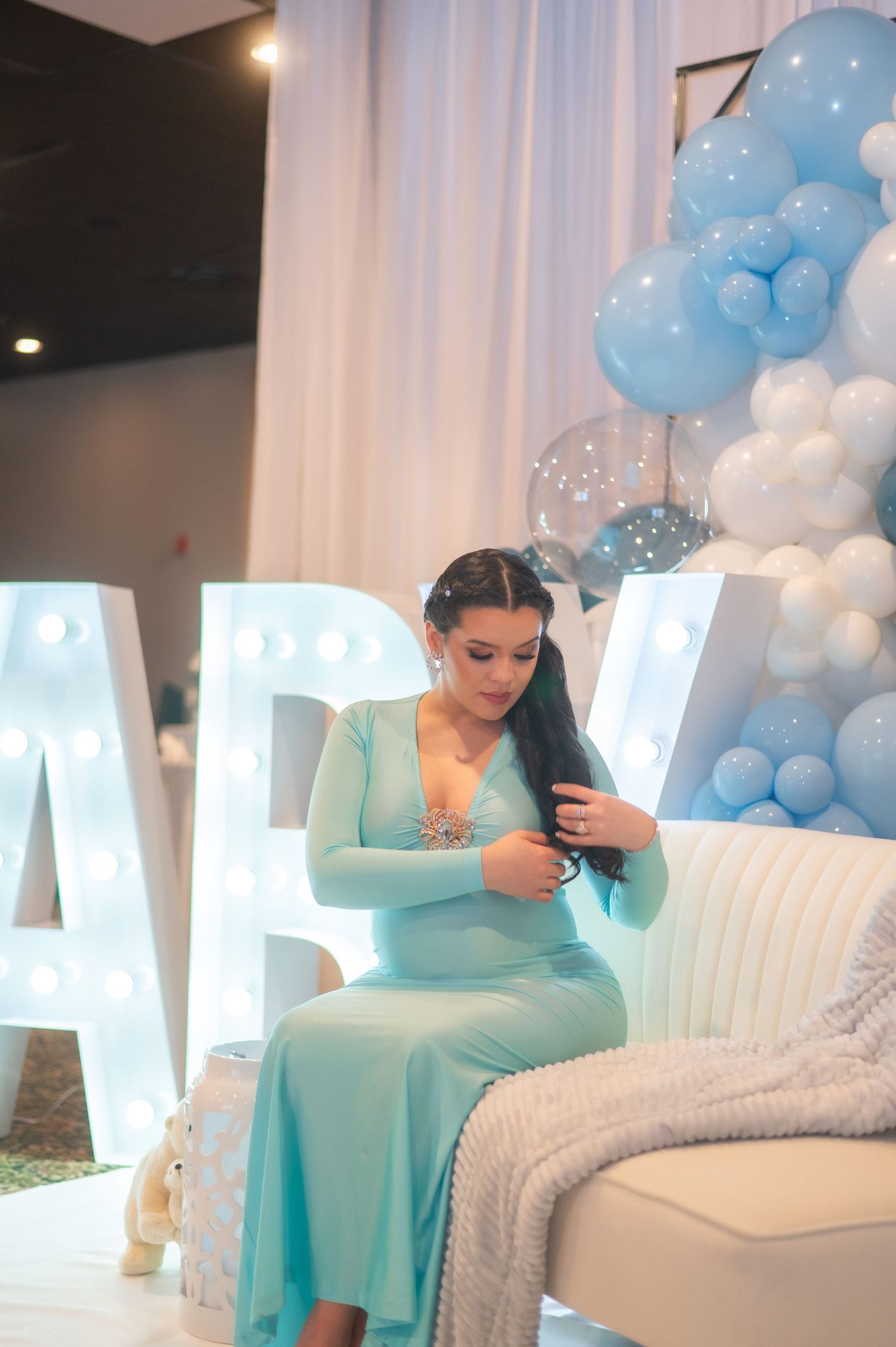 Woman in mint dress sits on white couch with illuminated letters and balloon decorations.
