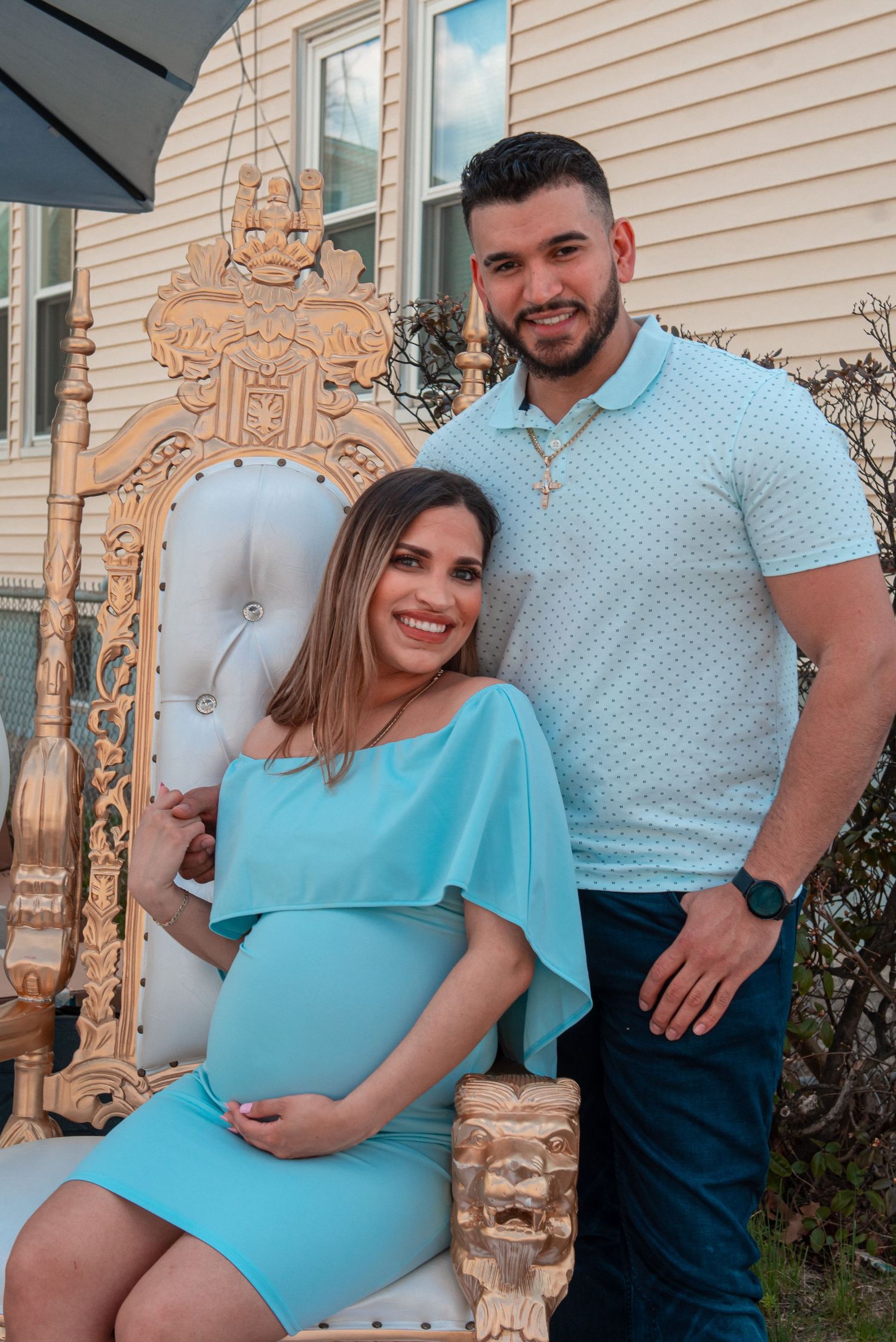 A couple poses together by an ornate gold throne chair during a maternity photo shoot.