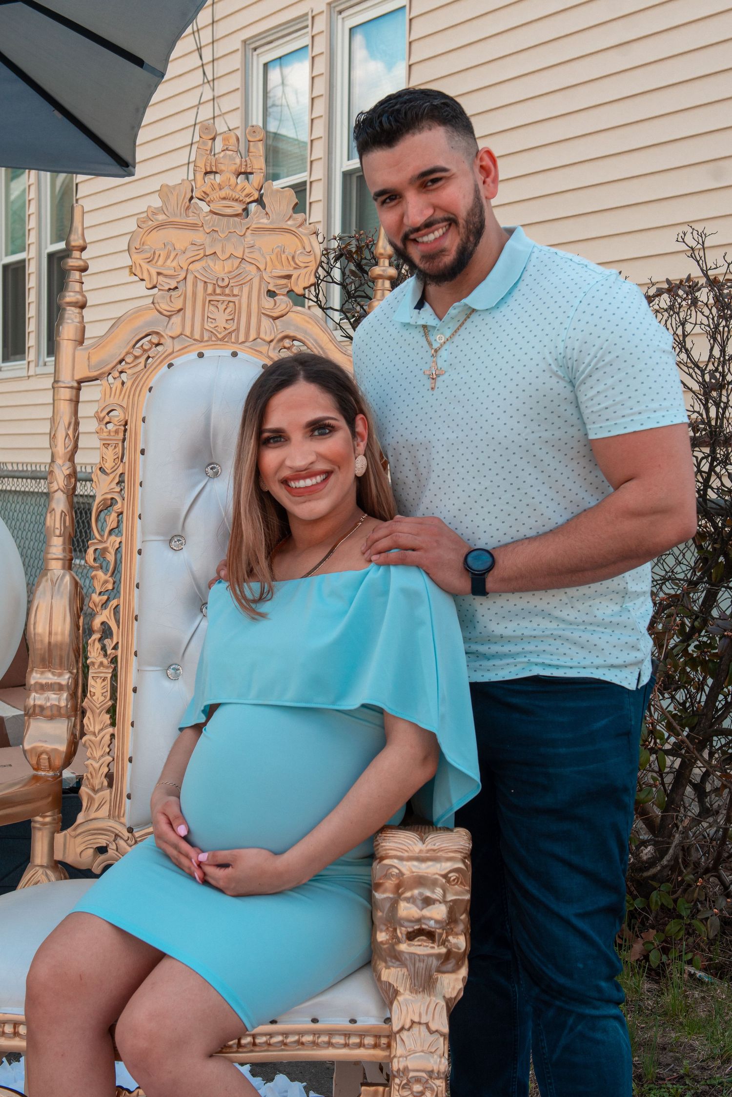 A couple smiles for a maternity photo against beige siding with a decorative gold throne.