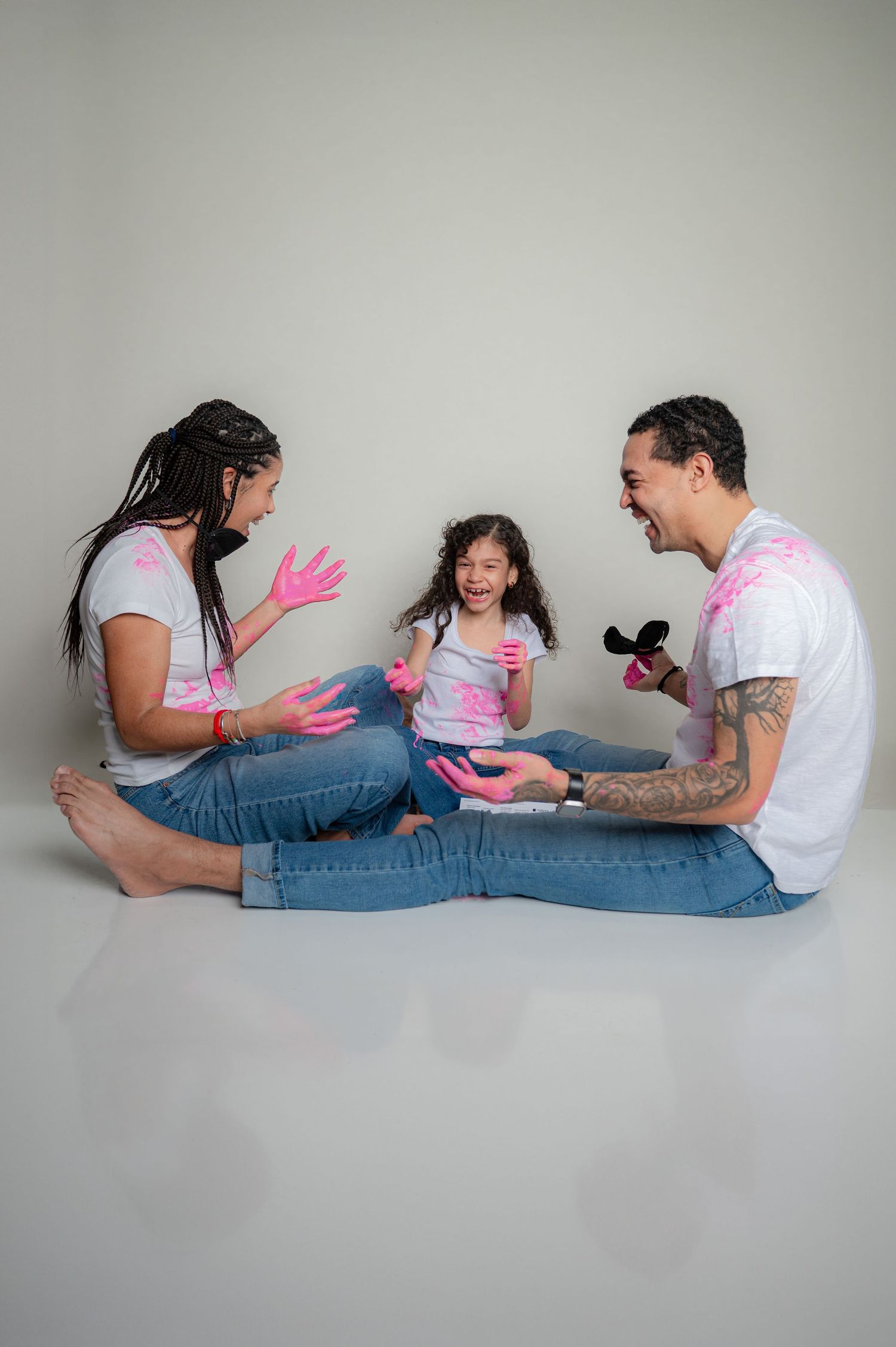 A family plays together on the floor wearing white shirts with pink paint splatter.
