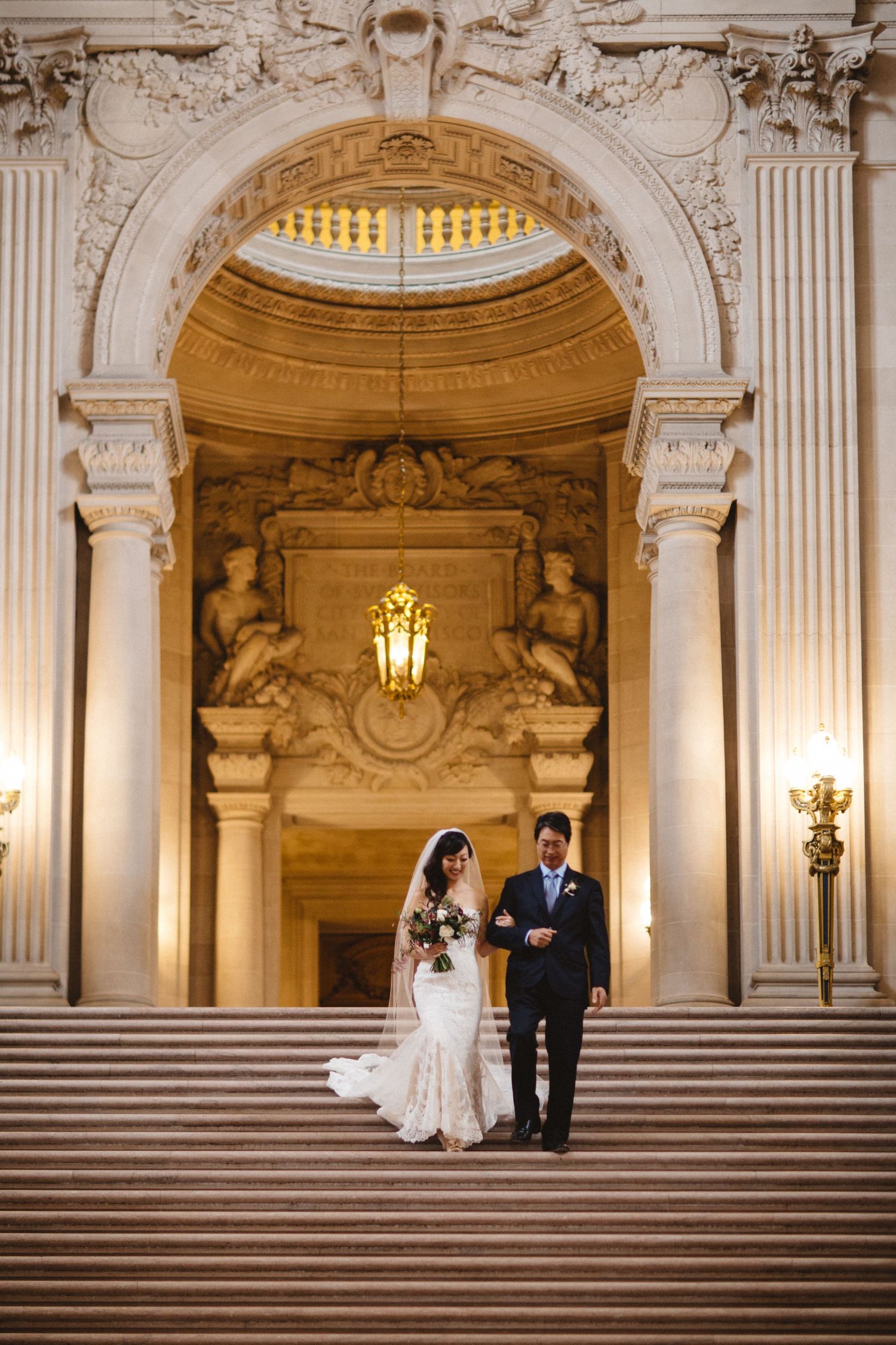 A wedding couple poses on marble steps beneath an illuminated classical architectural archway.