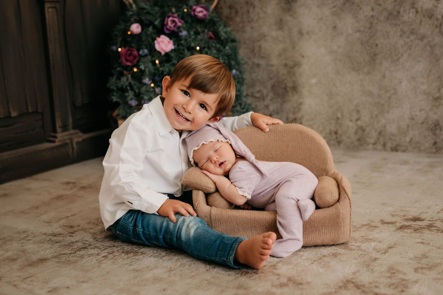 Fotografía de un hermanito sonriendo mientras posa con su hermana dormidita en un mini sofá en su sesión de fotos.