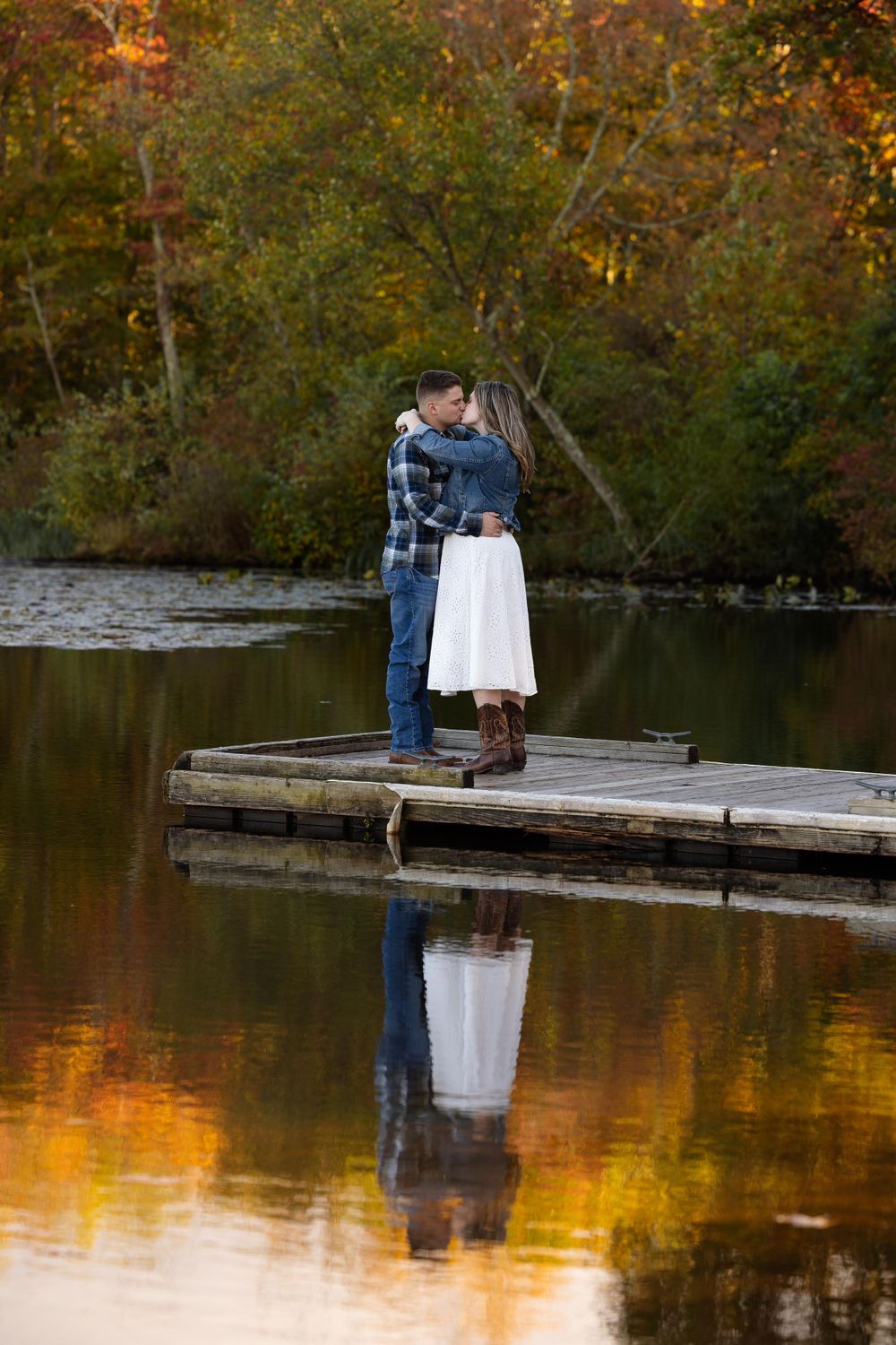 A couple embraces on a wooden dock over calm water reflecting golden autumn trees.