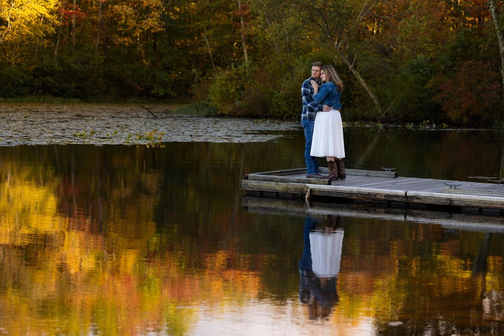 A couple stands on a wooden dock with golden autumn reflections in the still water.