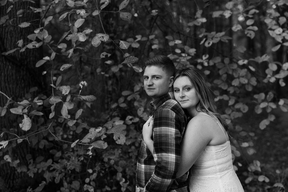 Romantic couple poses against leafy background in black and white portrait by trees.