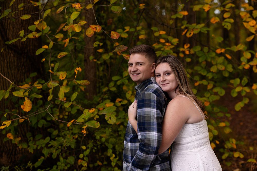 Couple embraces in front of autumn foliage wearing plaid shirt and white dress.