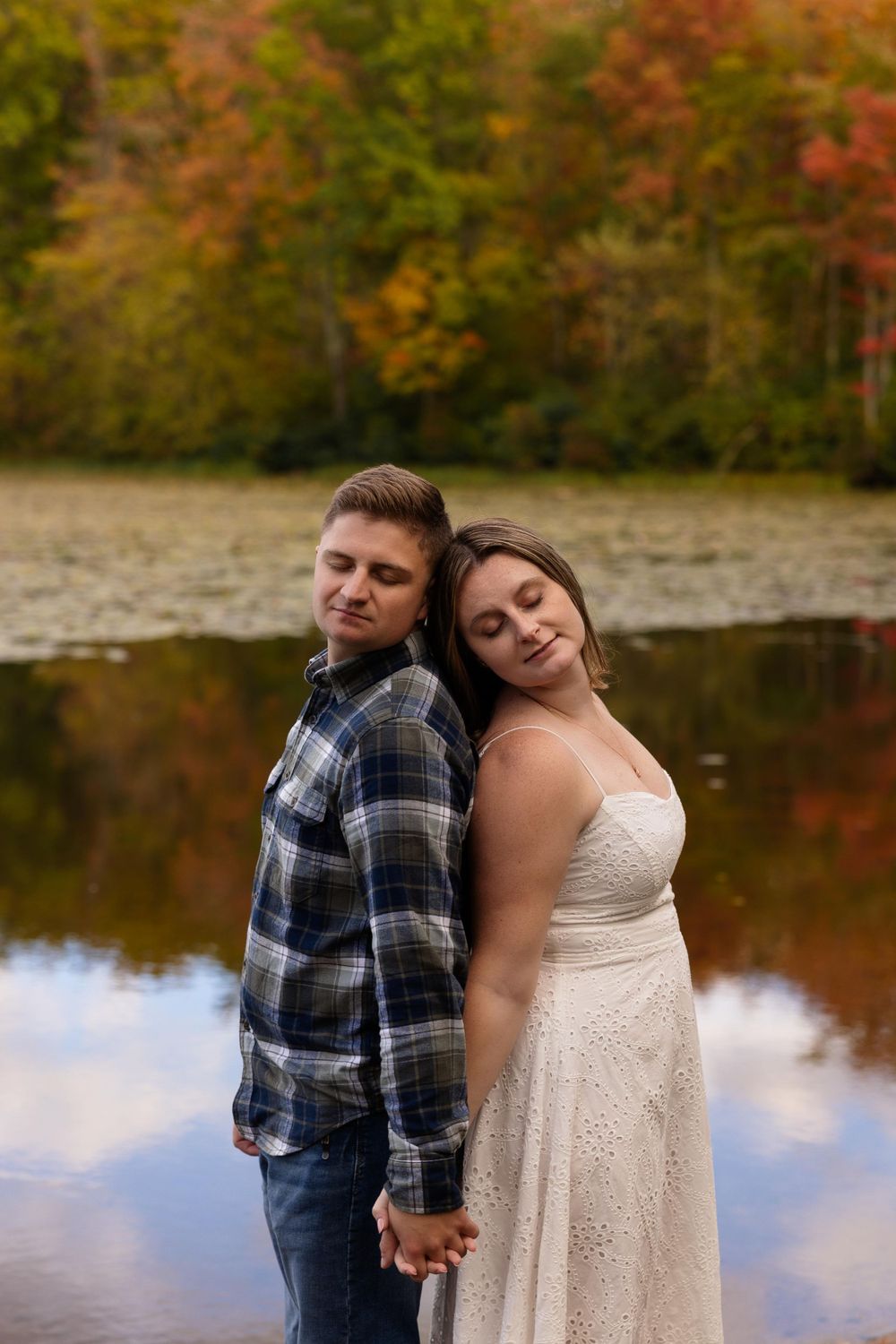Couple stands back-to-back by tranquil autumn pond with colorful foliage reflection.