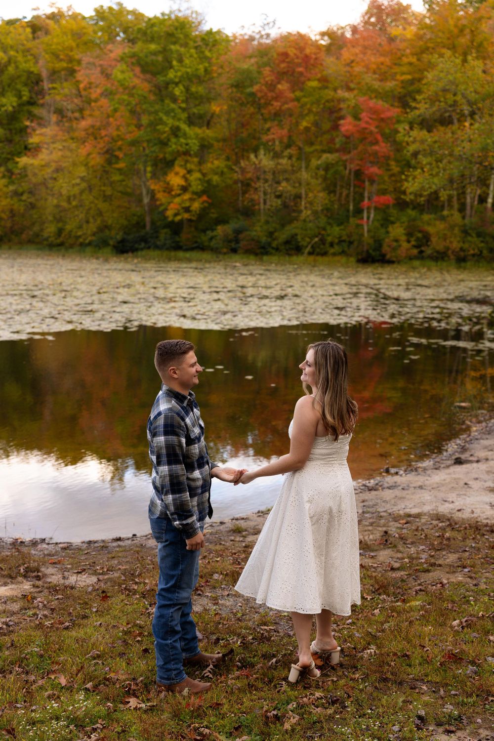 Couple holds hands lakeside with beautiful fall foliage reflecting in water.