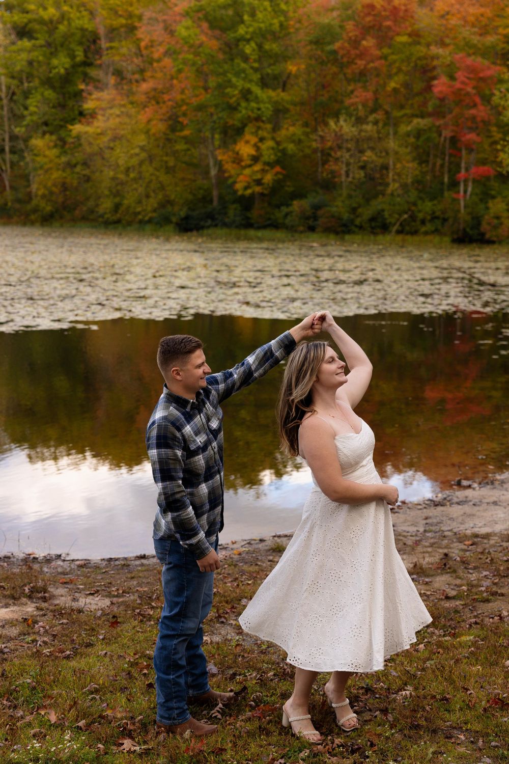 Dancing couple by autumn lake with partner twirling in white dress.