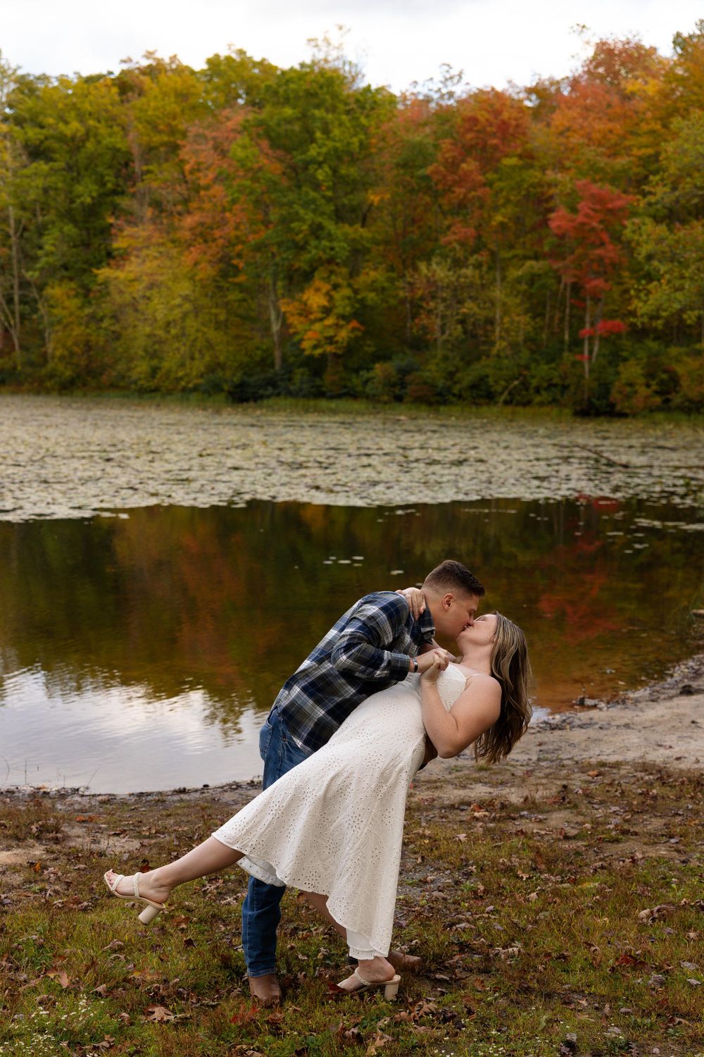 Romantic dip pose by autumn lake with colorful trees in background.