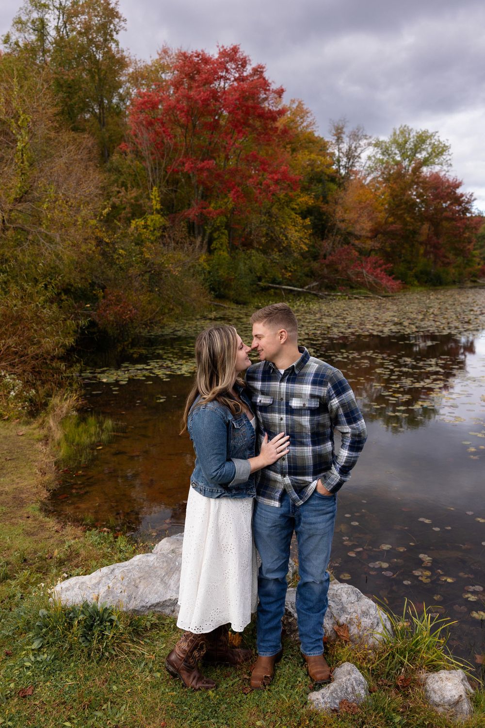 Couple standing by lake with vibrant fall foliage and reflecting water in background.