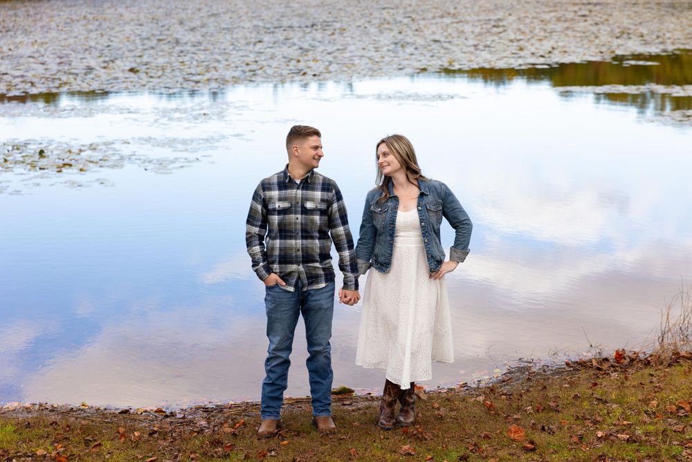 Couple standing by lakeside during fall engagement session with water reflections.