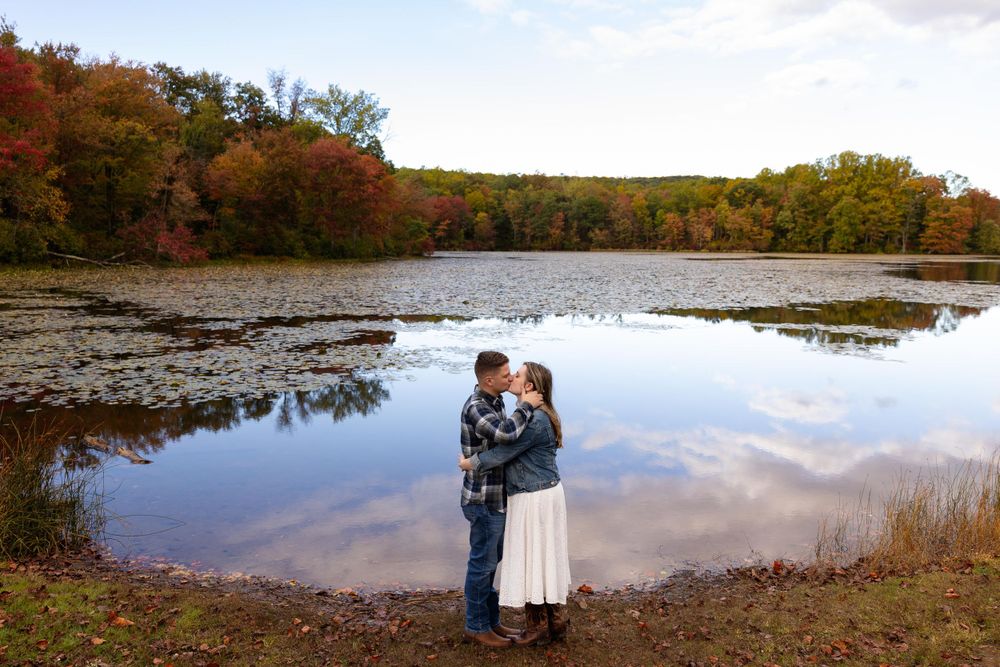 A couple in autumn attire embraces by a reflective pond surrounded by fall foliage.