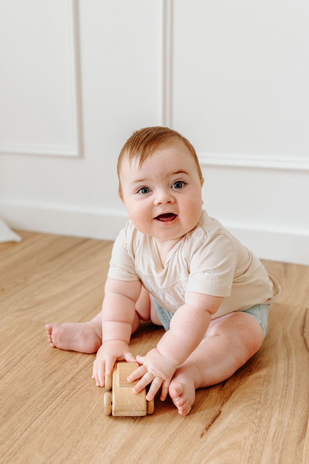 Baby in light colored outfit playing with wooden toy on hardwood floor.