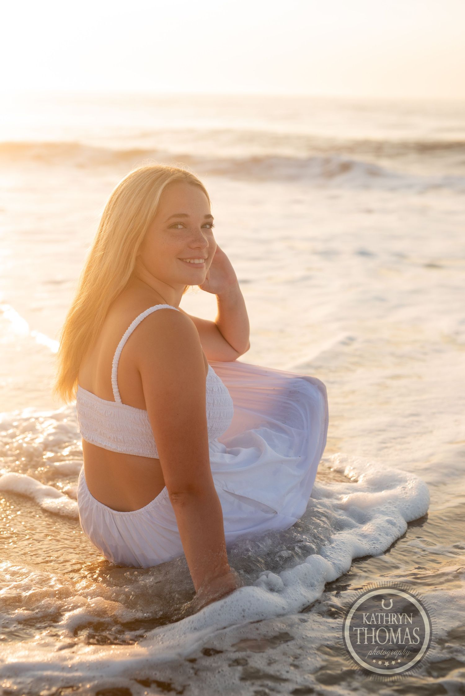 Person in white outfit sits at water's edge during sunset with waves rolling in.