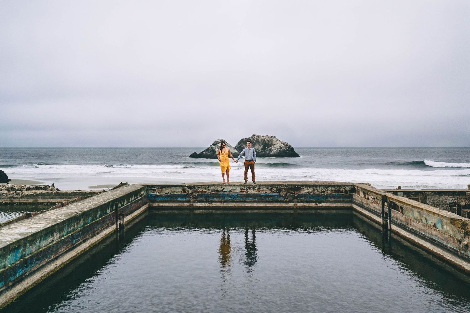 Two people stand on an oceanside pool structure with their reflection in the water below.