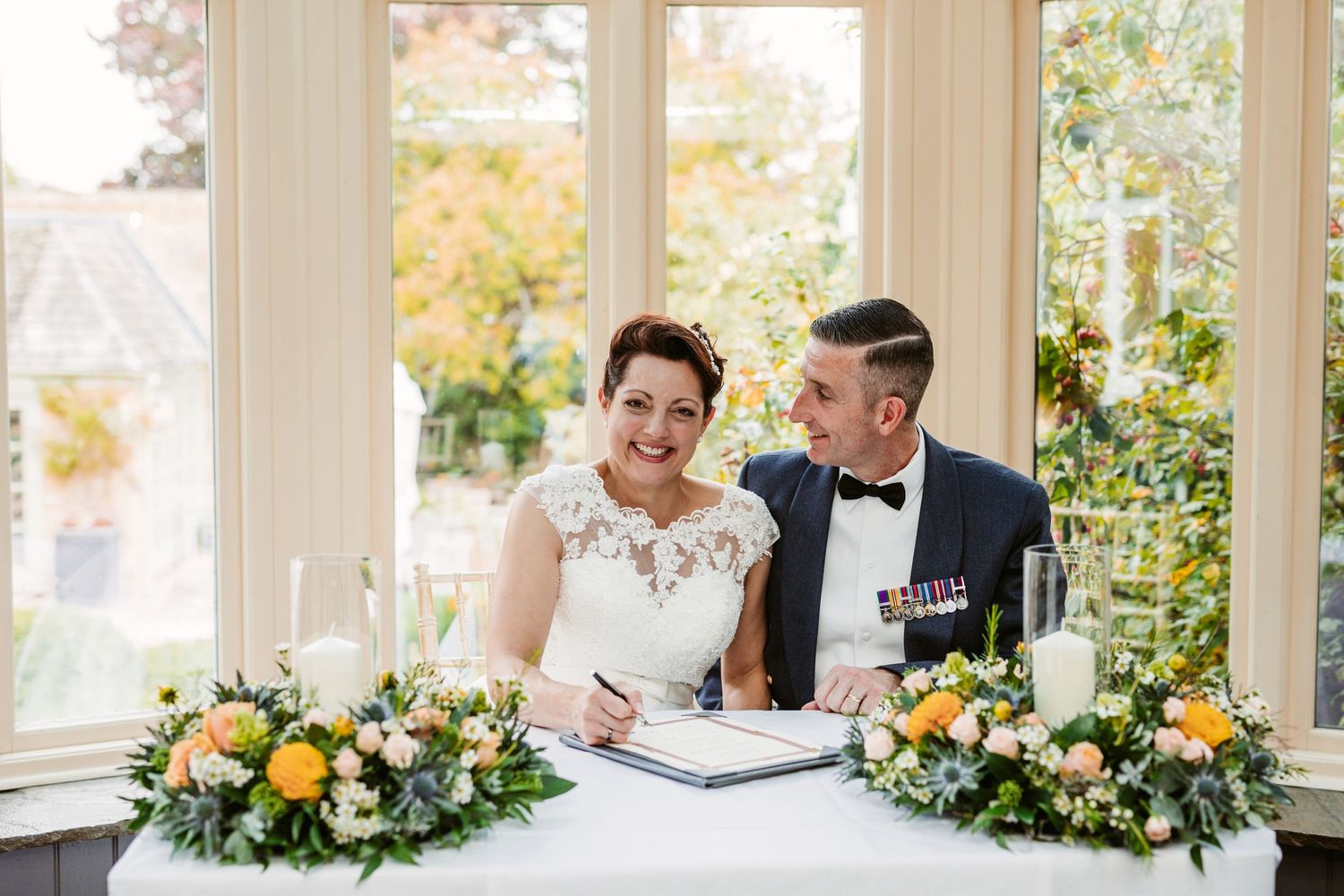 A wedding couple signs their marriage register beside beautiful yellow and white floral arrangements.