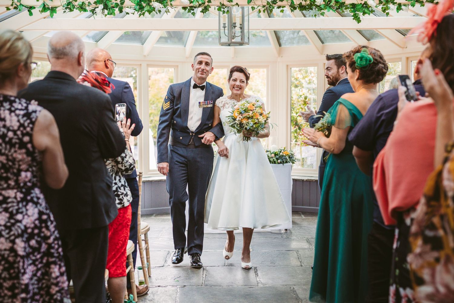 Wedding guests line the aisle during a beautiful ceremony in a glass-roofed venue.