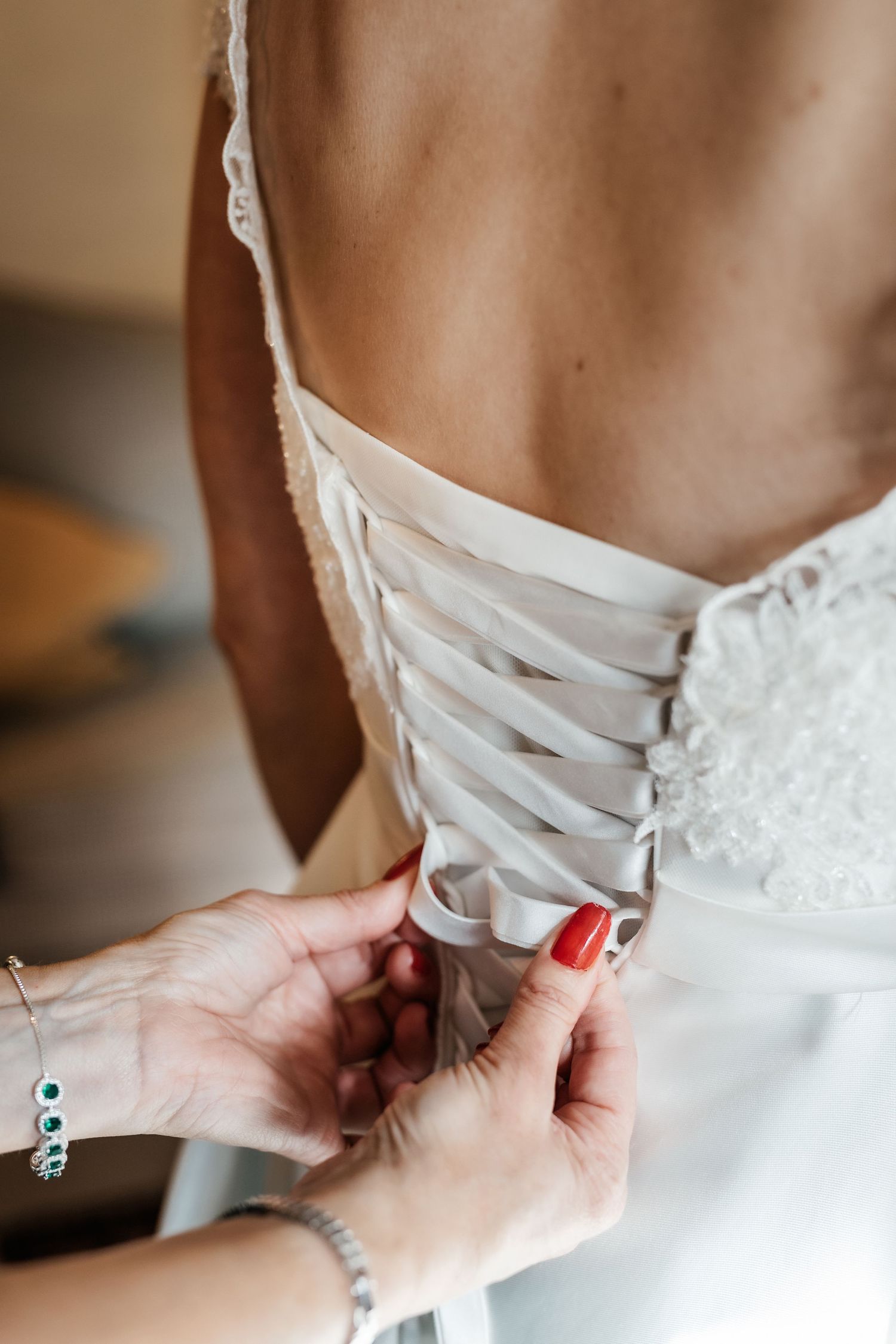 Close-up of hands lacing up a white wedding dress corset with delicate ribbons and red nail polish.