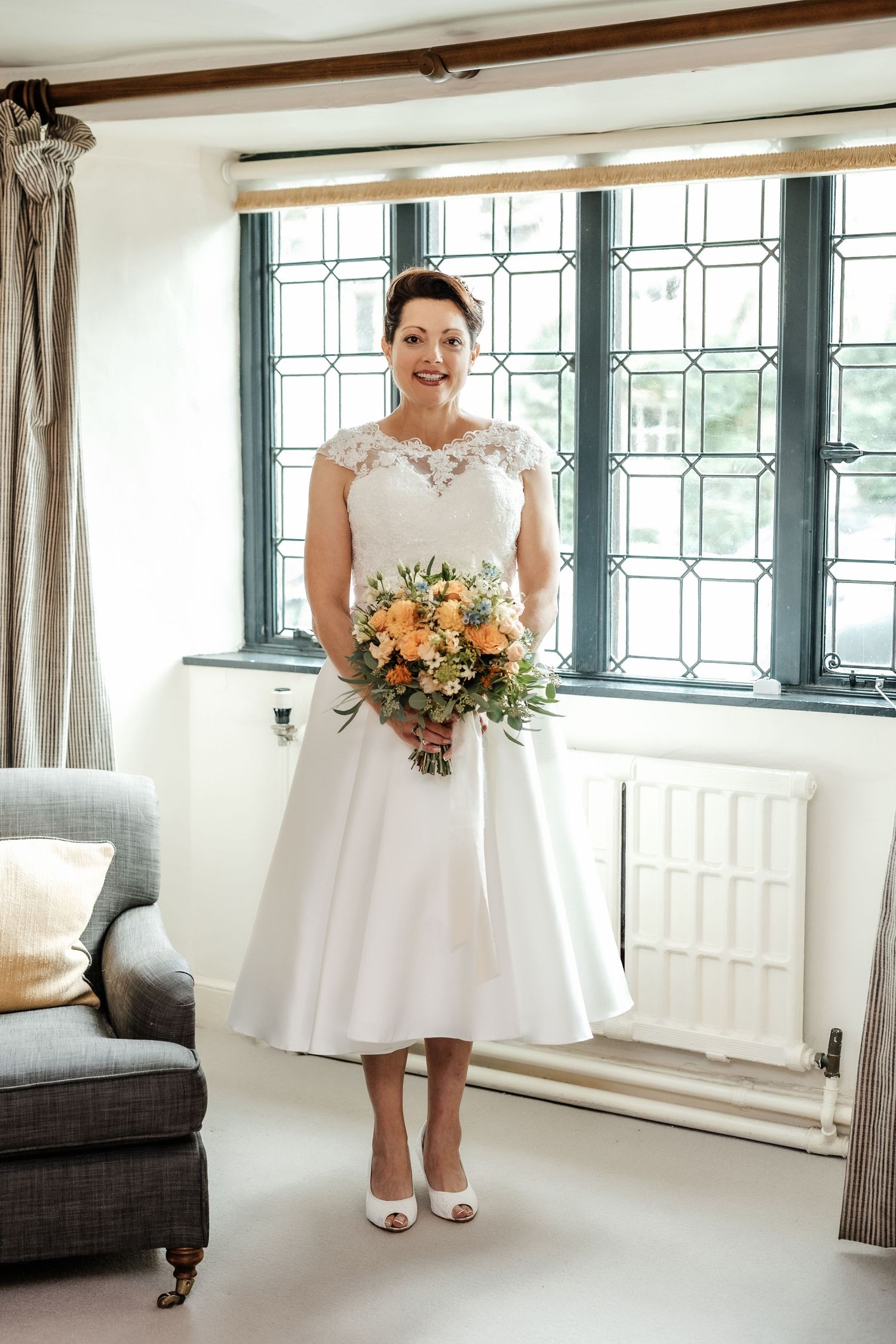 Tea-length wedding dress with lace details and peach bouquet against a window backdrop.