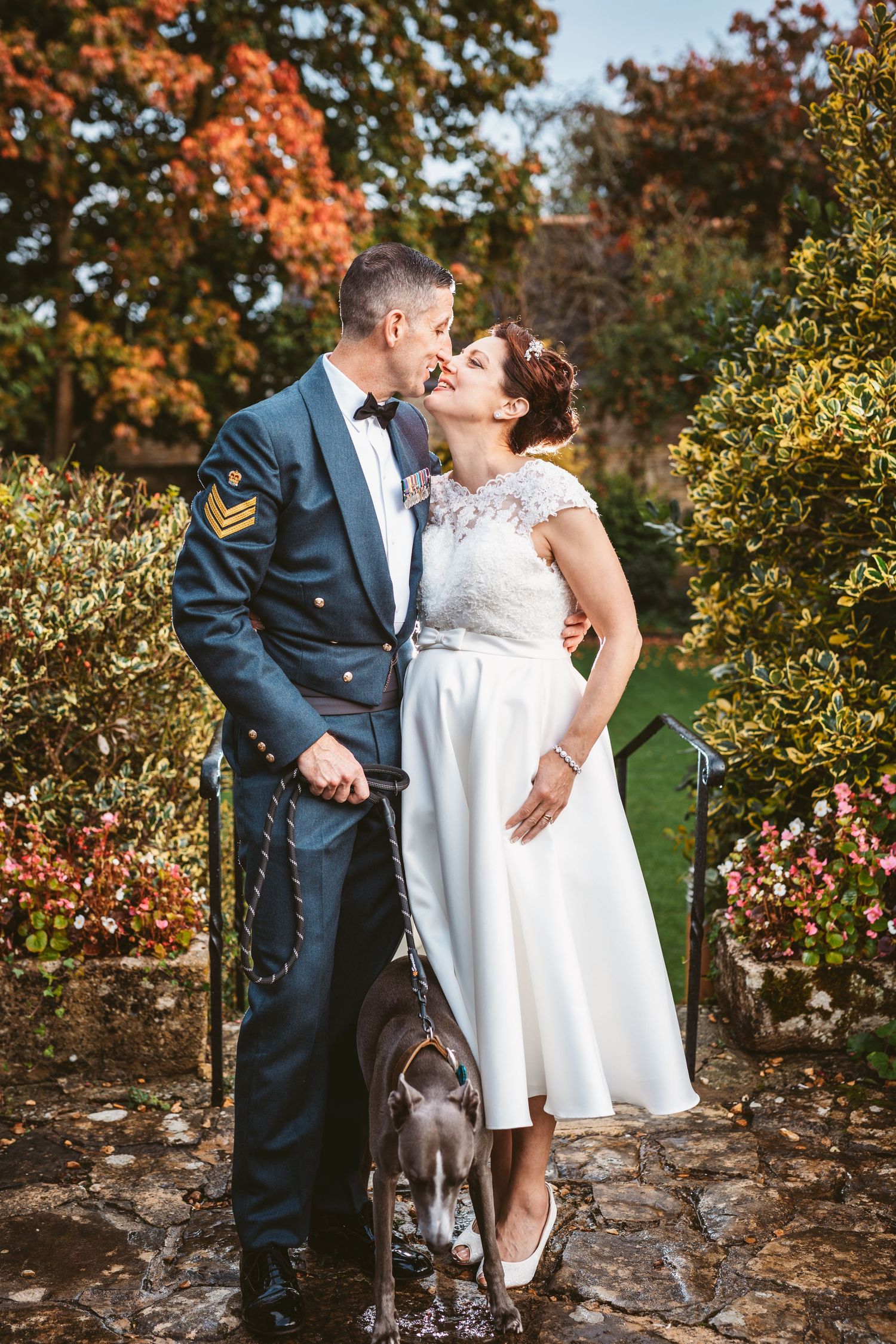 Military groom and bride in tea length dress posing with their pet dog in autumn garden setting.