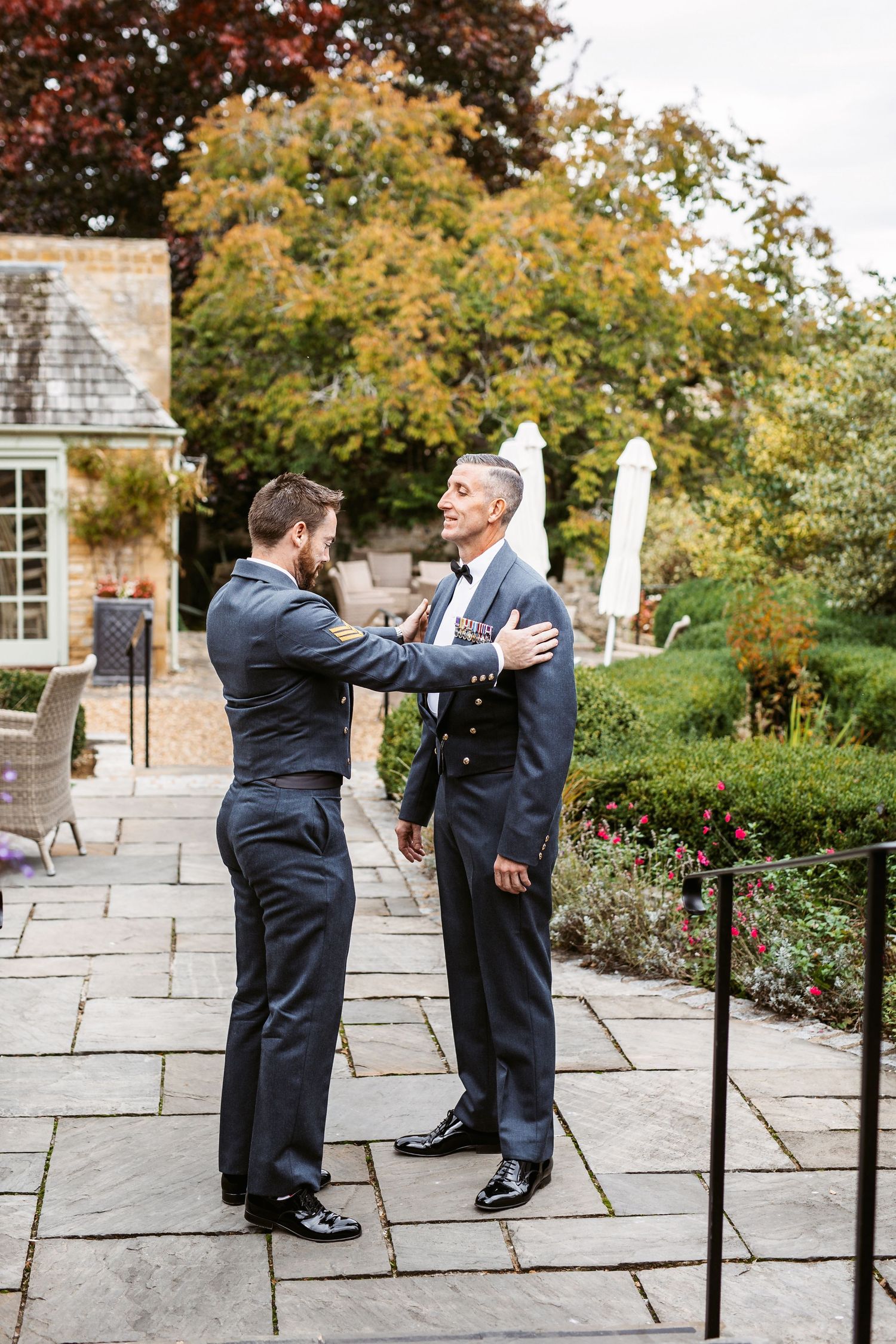 Two people adjusting formal attire on stone patio with fall foliage in background.