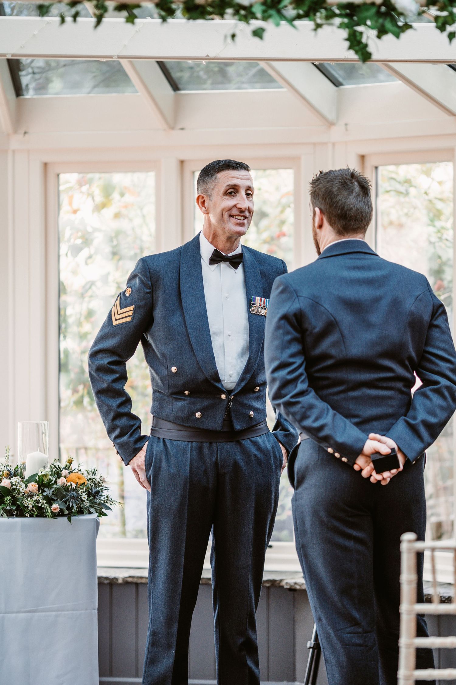 Two men in formal military dress uniforms having a conversation in a conservatory setting.