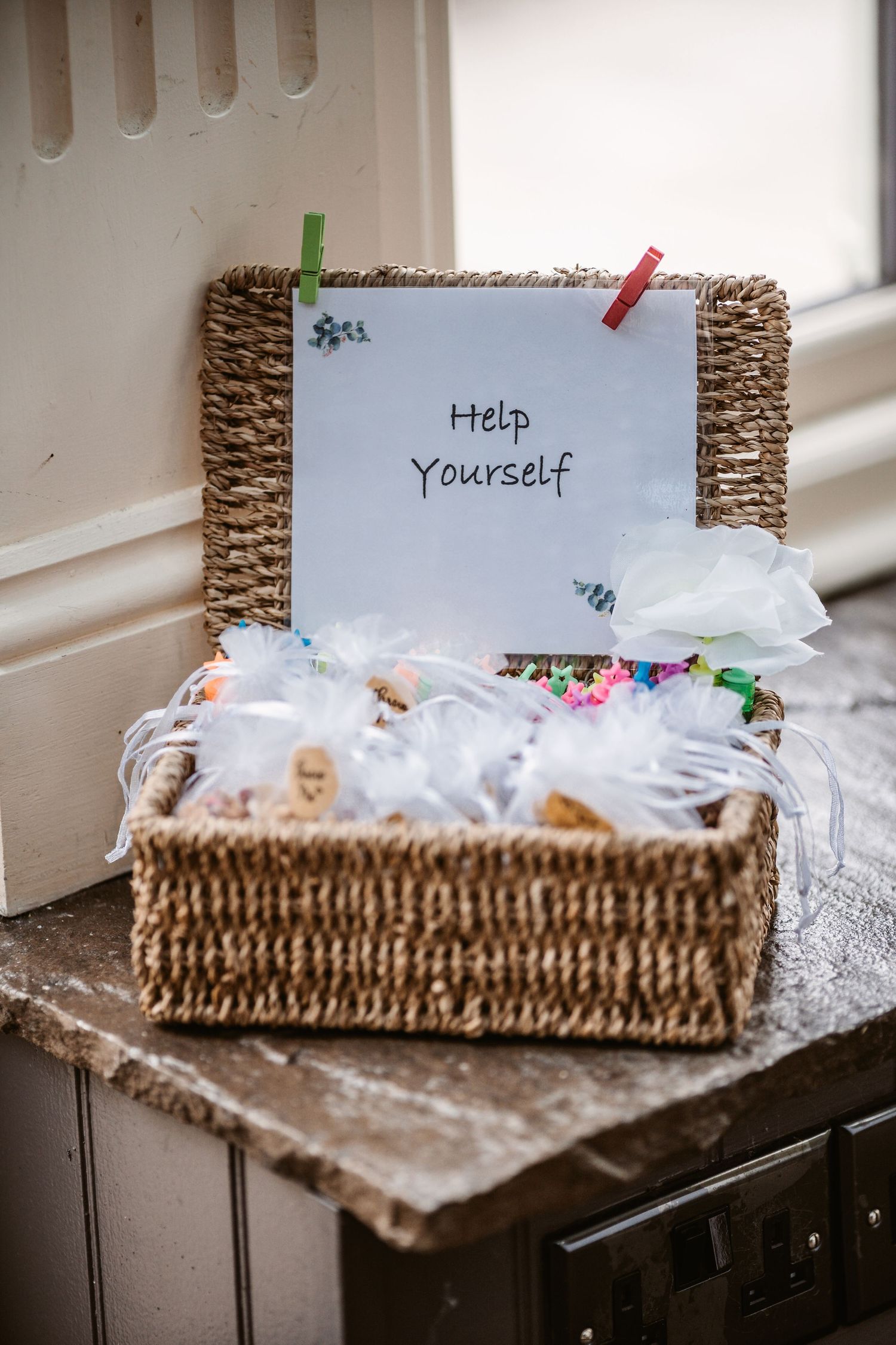 A woven basket filled with wedding favors and a 'Help Yourself' sign attached with colorful clips.
