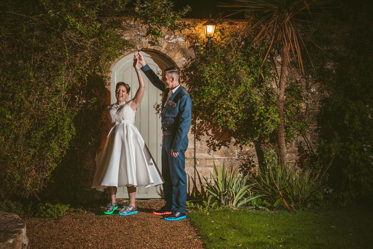 A couple strikes a fun pose with raised arms under a garden archway at night.