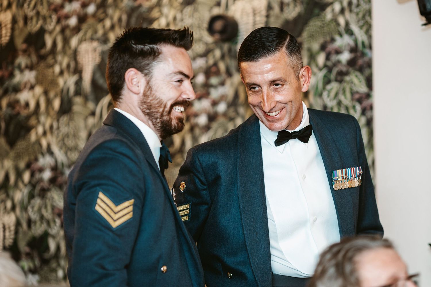 Two servicemen in military dress uniforms sharing a laugh at formal event.