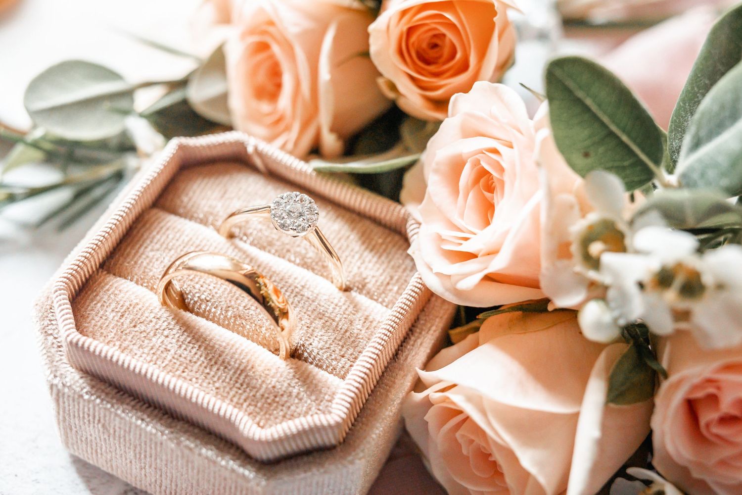 Wedding rings nestled in pink velvet box with peach roses and eucalyptus leaves.