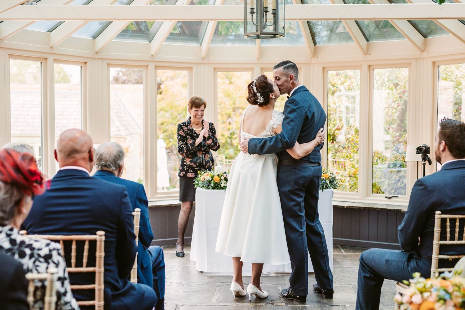 An intimate wedding ceremony taking place in a sunlit conservatory with guests seated on white chairs.