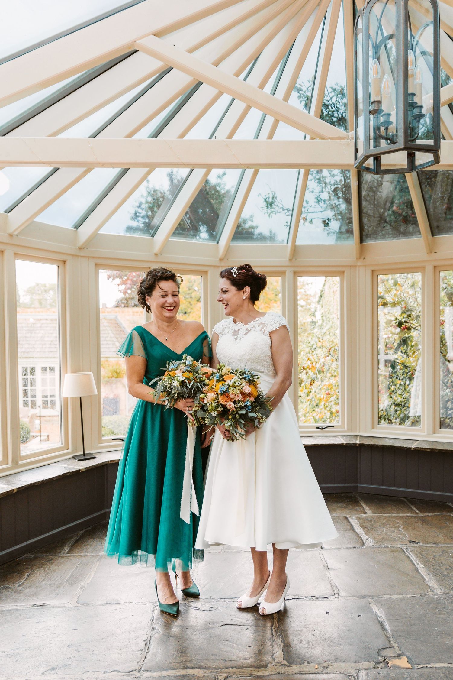 A wedding dress and emerald green gown stand together in a sunlit conservatory.