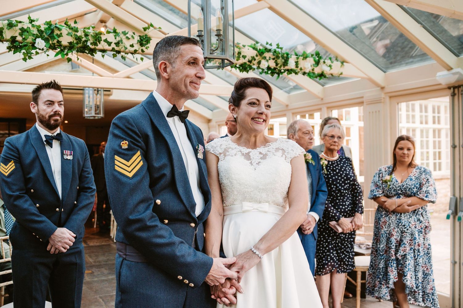 Ceremony in a glass-roofed venue with greenery garlands and formal military attire.