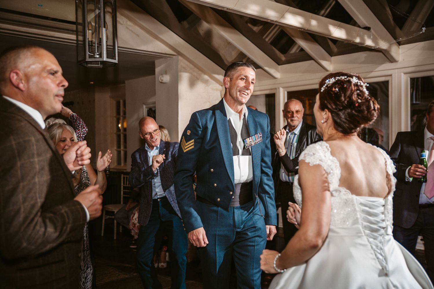 Wedding guests surround a dancing couple at an indoor reception with rustic wooden beam ceiling.