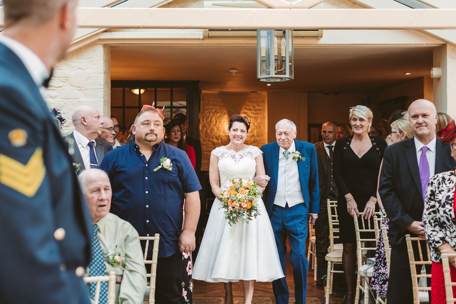 Wedding ceremony in a glass conservatory with guests seated and floral decorations.