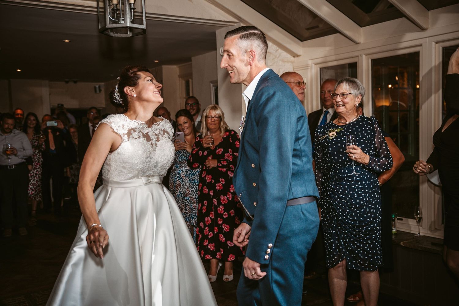 Wedding couple laughs together during their first dance while guests look on at an indoor reception.