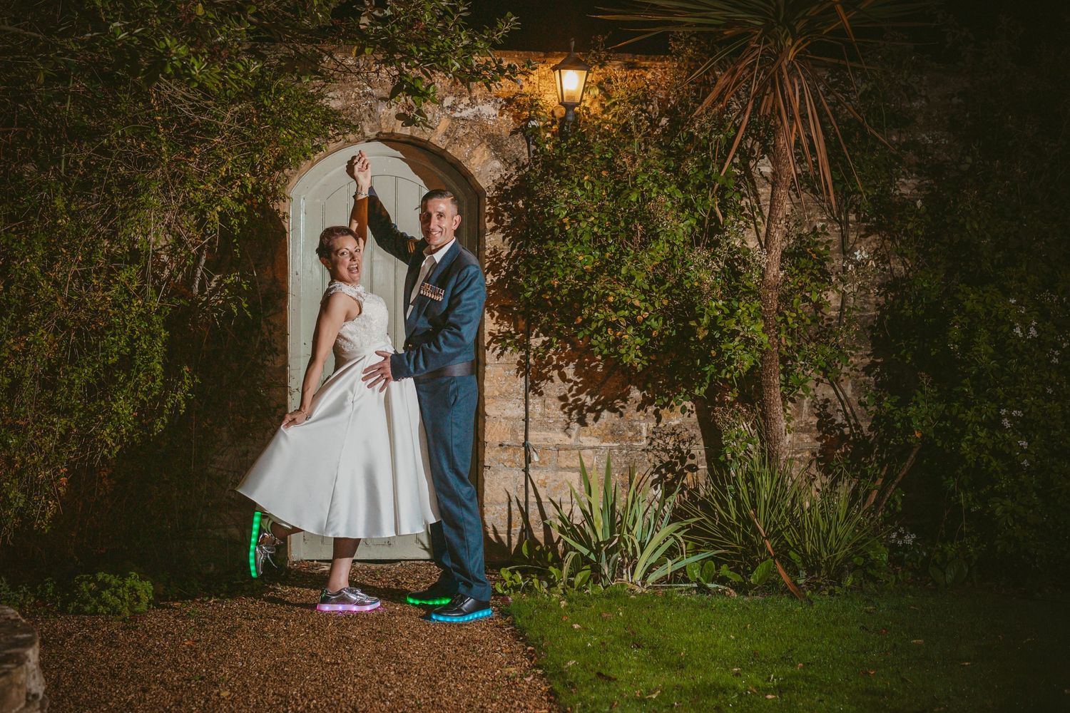 A couple poses elegantly beneath a lit garden archway with greenery surrounding them.