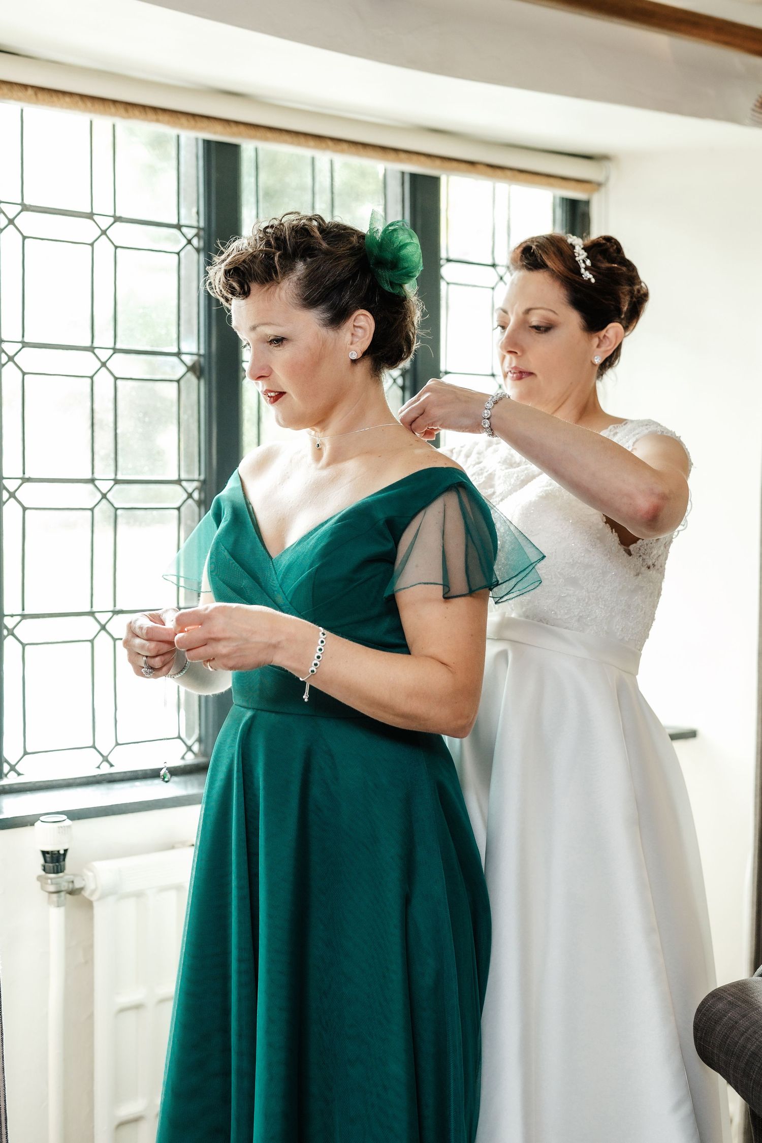 Two wedding attendees in a sunlit room, one wearing emerald green and one in white.