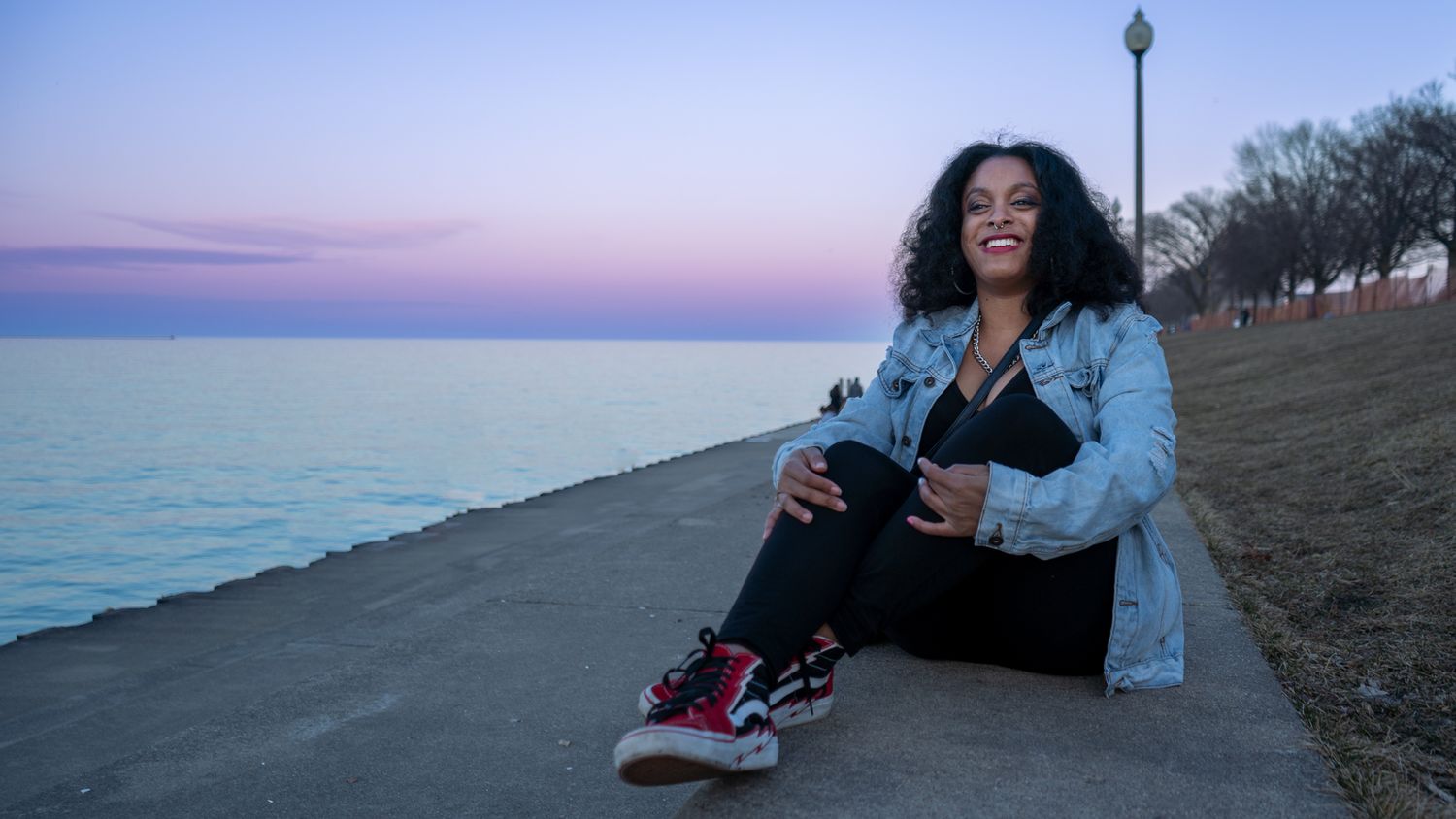 Person relaxing by lakefront at sunset wearing denim jacket.