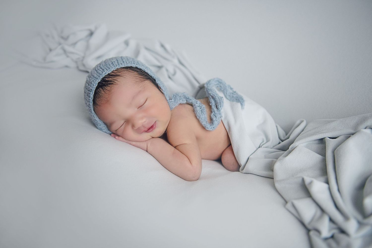 Sleeping newborn wearing blue bonnet and wrapped in soft grey fabric during portrait session.