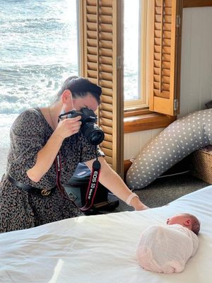 Photographer capturing a newborn baby photo session with ocean waves visible through window shutters.