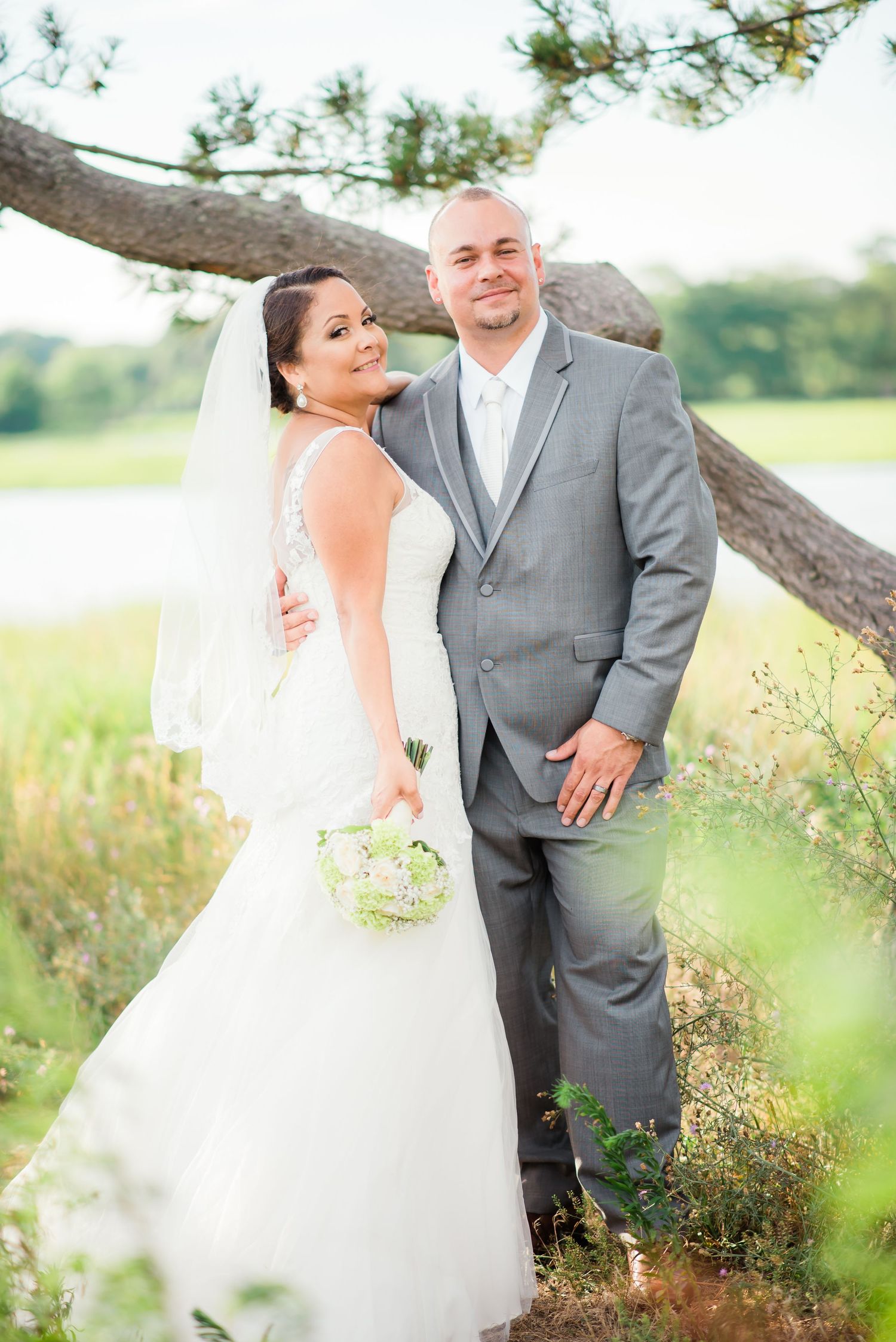 Newlyweds pose together by a tree in a sunlit field with the bride holding a bouquet.