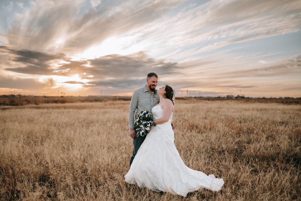 Wedding couple embraces in golden field during dramatic sunset with wispy clouds overhead.