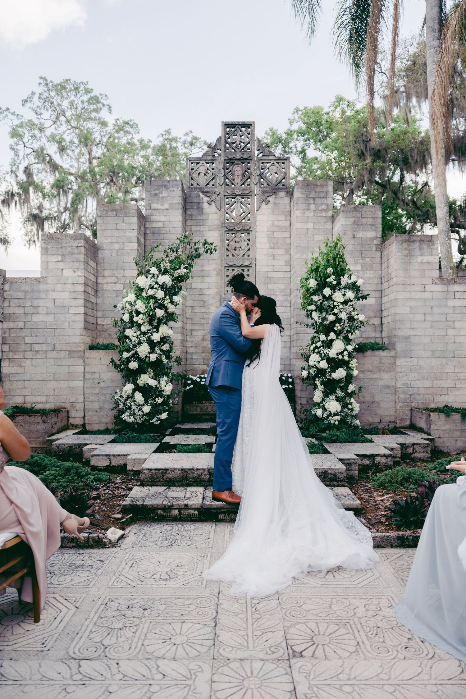 A wedding ceremony scene with floral arches and a stone wall backdrop.