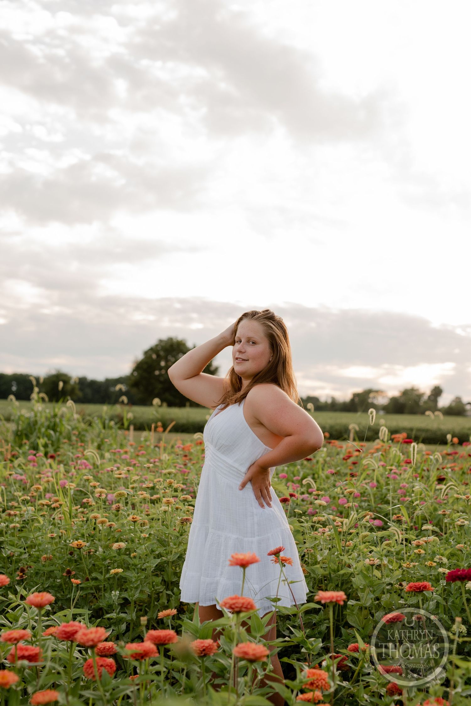 Person in white dress poses in field of orange and pink wildflowers at sunset.