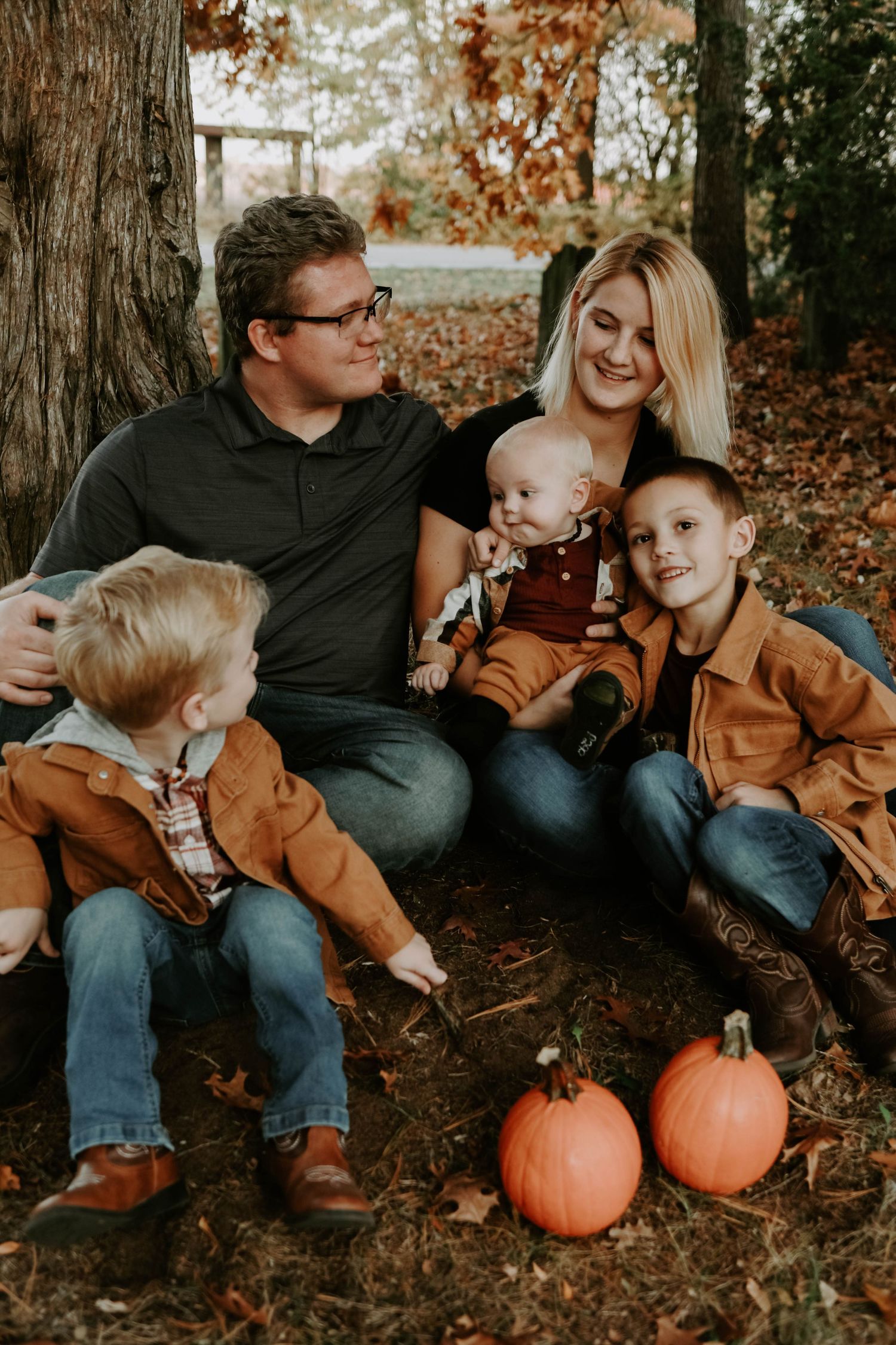 Family sits together on fallen leaves with orange pumpkins during autumn.