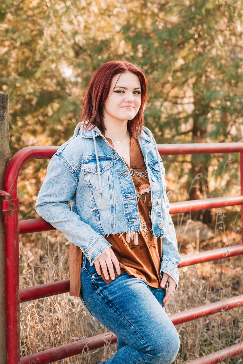 A woman in denim jacket leans against red metal gate in rustic outdoor setting.