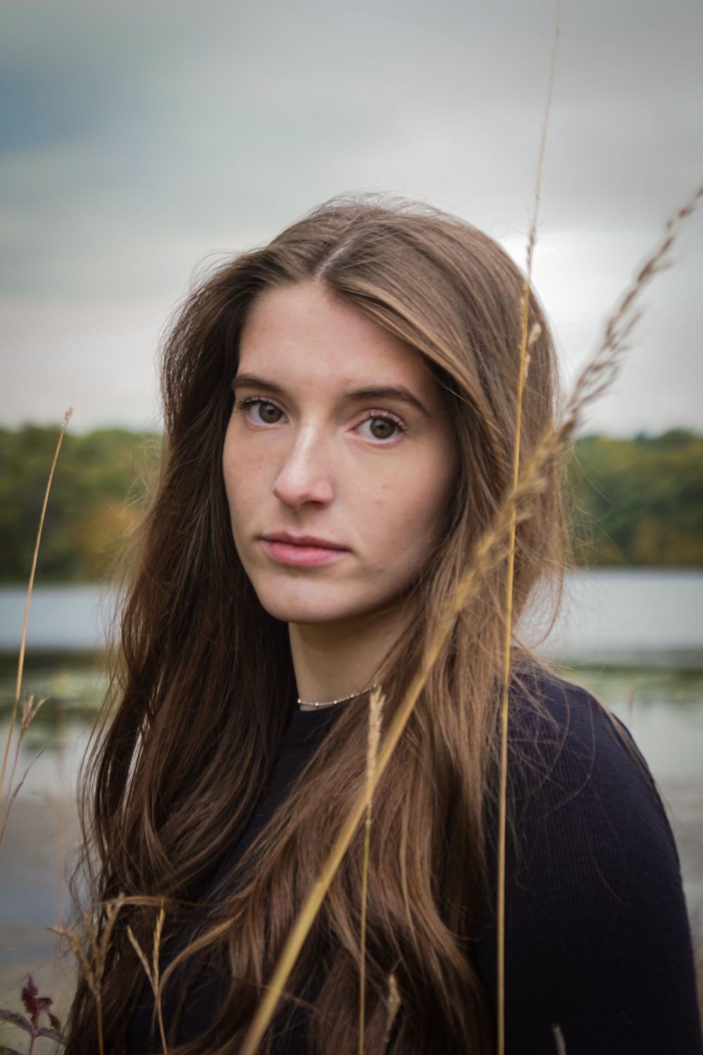 Portrait of a young woman with long brown hair against a lakeside background with tall grass in soft lighting.