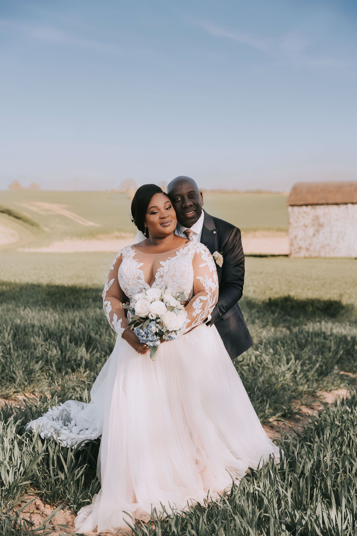 A romantic wedding photoshoot in a lush green field with the couple embracing and holding a white bouquet.